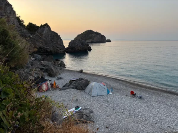 Chiliadou Beach in Euboea, Greece, features serene waters, rocky cliffs, and scattered tents along the pebbled shoreline at sunset.