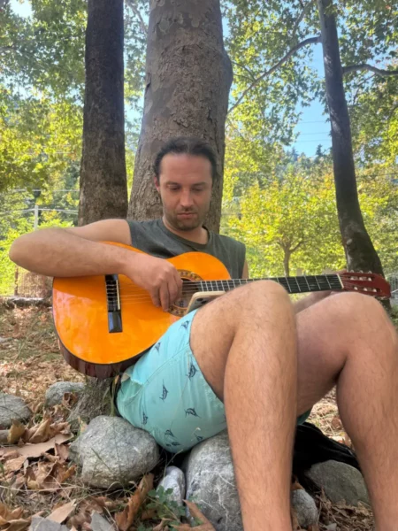A man sits on rocks by a tree at Chiliadou Beach, playing an orange guitar in a lush green setting on Euboea, Greece.