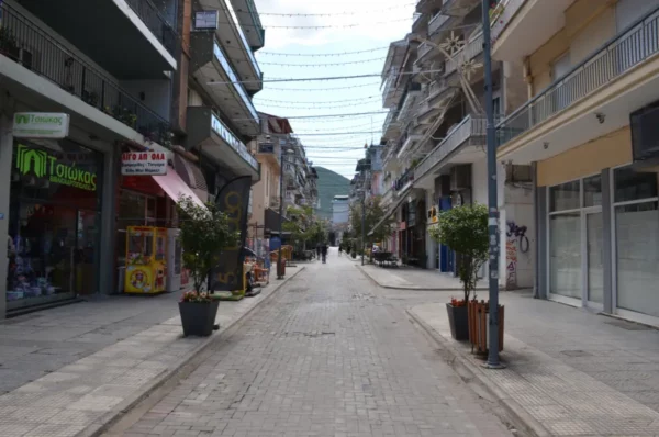 Quiet street scene in Florina, Greece, featuring shops, trees lining the walkway, and distant mountains under a clear sky.
