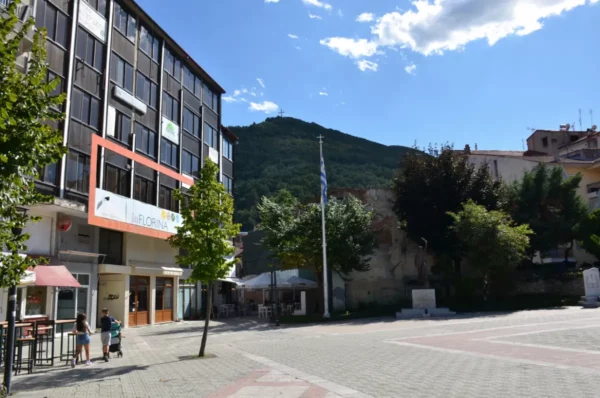 Bright square in Florina, Greece, featuring modern buildings, a flagpole, and mountainous backdrop under a clear blue sky.