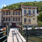 Pedestrian bridge over the Sakoulevas River in Florina, with neoclassical houses along the riverbank and a green hillside in the background.