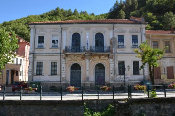 Historic building with an ornate façade in Florina, Greece, framed by vibrant greenery and colorful flowers along the street.