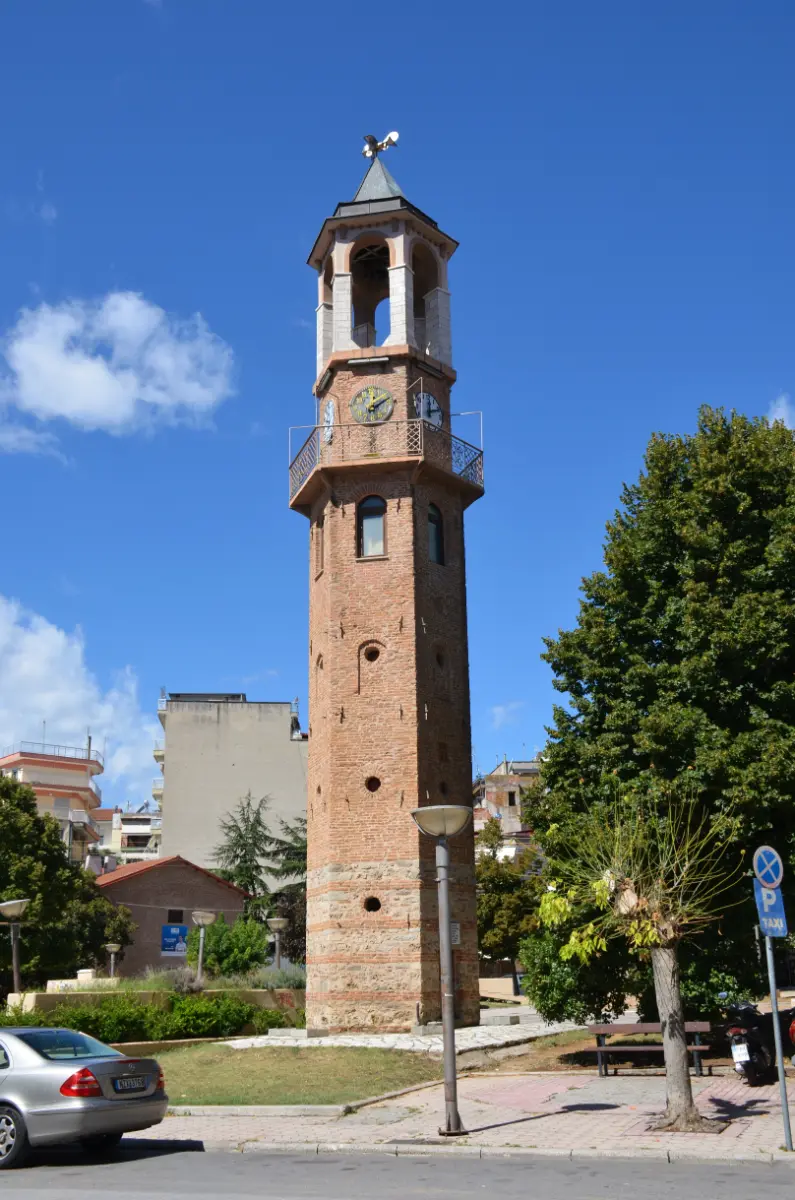 The historic clocktower of Grevena, Greece, rising above a small square with trees and low buildings under a clear blue sky.
