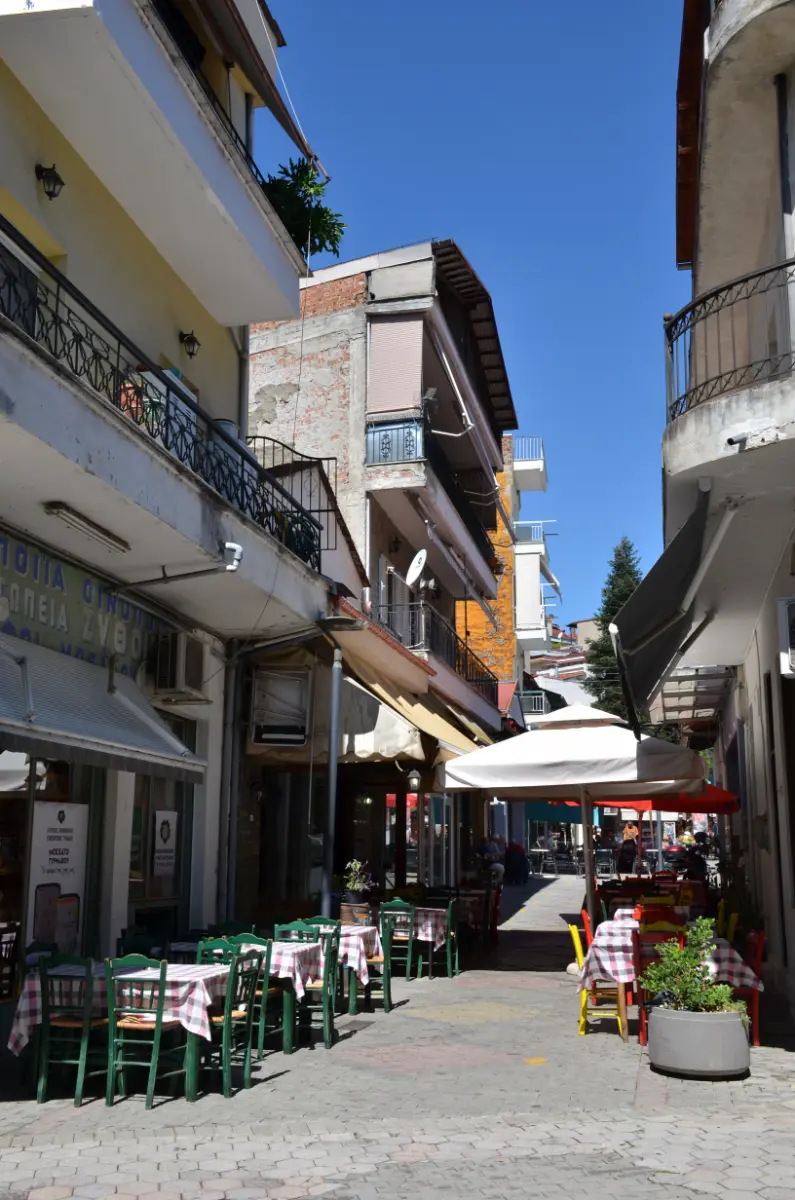 Narrow pedestrian street in Grevena, Greece, lined with tavern tables, green chairs, and cafés under umbrellas on a sunny day.