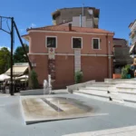 Emilian Square in Grevena, Greece, with curved amphitheater-style steps, a small fountain, and surrounding cafés on a sunny day.
