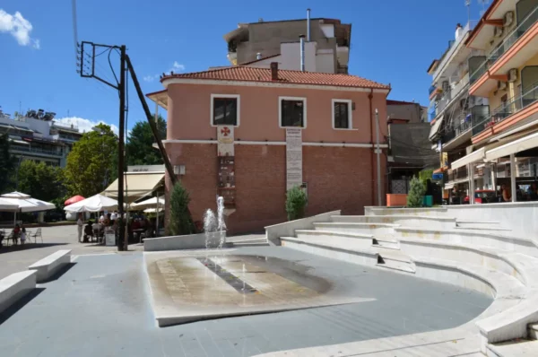 Emilian Square in Grevena, Greece, with curved amphitheater-style steps, a small fountain, and surrounding cafés on a sunny day.