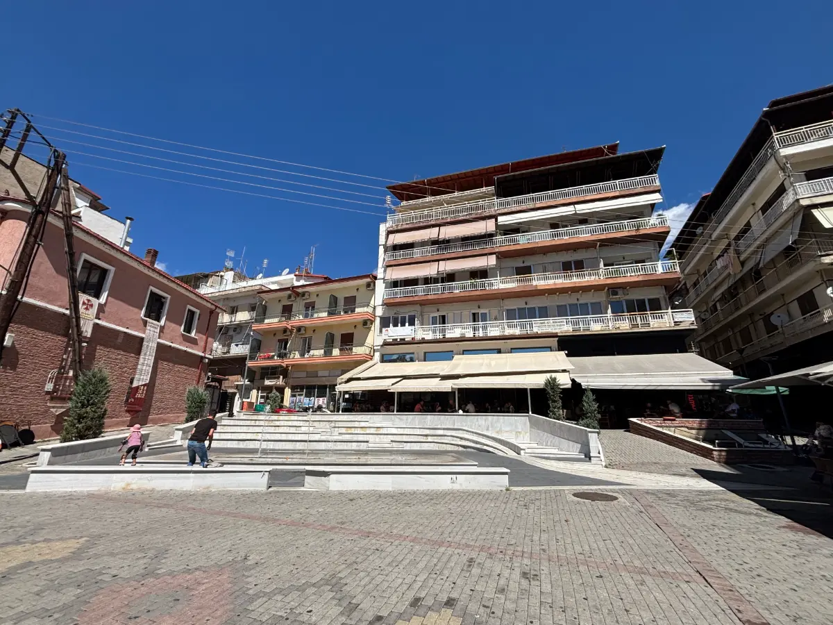 Emilian Square in Grevena, Greece, with amphitheater-style steps, a central fountain, surrounding cafés, and apartment buildings under a clear sky.