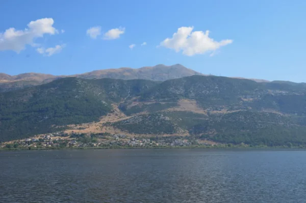 Stunning view of the tranquil lake surrounded by mountains near Ioannina, Greece, under a clear blue sky with fluffy clouds.