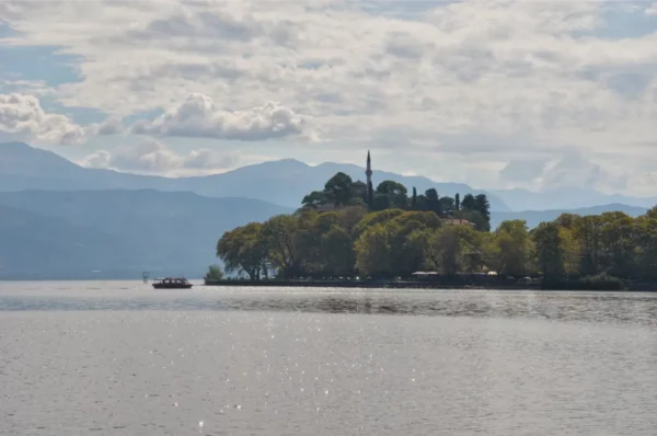 Idyllic lake view in Ioannina, Greece, with a boat cruising by against a backdrop of mountains and lush greenery.