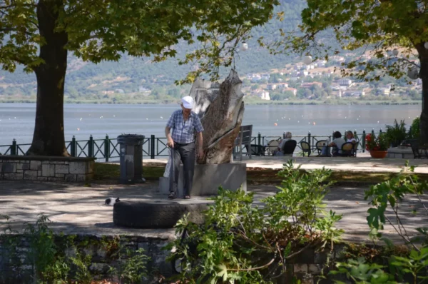 Man tending to a monument by the lake in Ioannina, Greece, surrounded by trees and a peaceful waterfront scene.