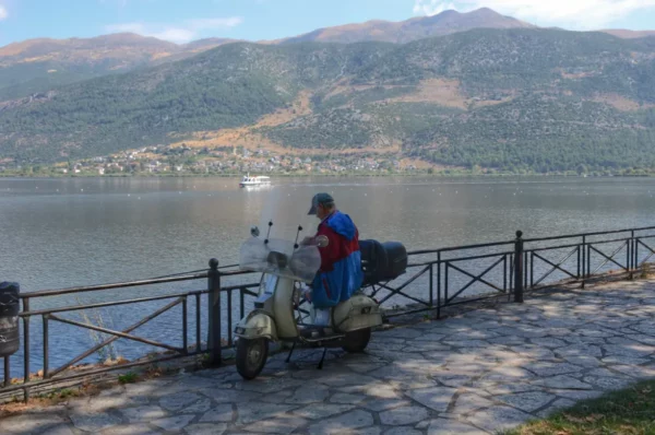 A man in a blue raincoat sits on a scooter by the lake, framed by mountains and clear skies in Ioannina, Greece.
