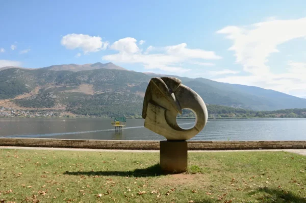 Sculptural artwork beside the lake in Ioannina, Greece, with mountains in the background and a clear blue sky.