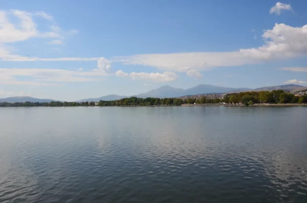Serene waters reflect the sky and mountains near Ioannina, Greece, surrounded by lush greenery along the shore.