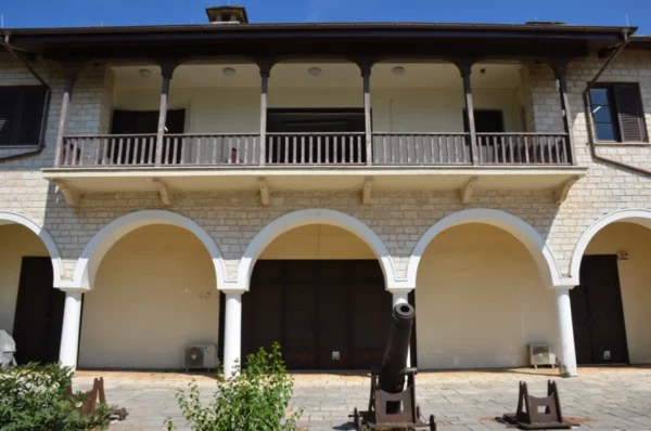 Historic stone building with arched entrances and a cannon in front, nestled in the scenic city of Ioannina, Greece.
