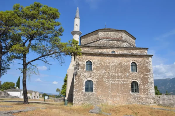 Historic stone mosque stands under a clear blue sky in Ioannina, Greece, surrounded by lush greenery and arid terrain.