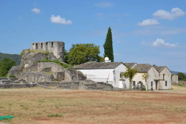 Historic ruins and a rustic building overlook a dry landscape in Ioannina, Greece, under a clear blue sky.