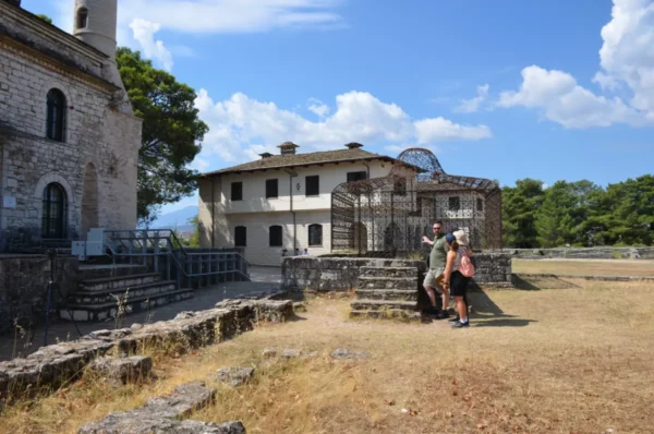 Visitors enjoy a sunny day near historic stone structures in Ioannina, Greece, with clouds and trees in the background.