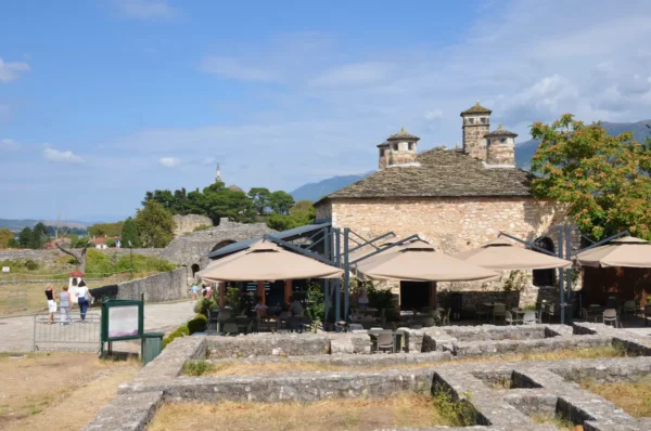 Historic stone building with a slate roof overlooks outdoor cafes in Ioannina, Greece, set against a clear blue sky and lush greenery.