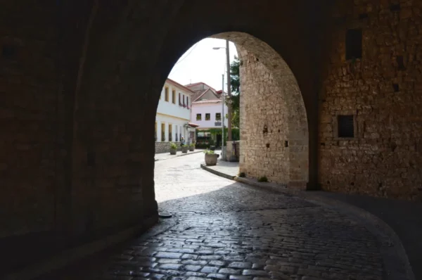 Stone archway leading to a sunny street in Ioannina, Greece, with charming buildings and cobblestone paving visible.