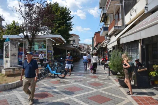 Lively street scene in Ioannina, Greece, featuring shops, pedestrians, and a bike rental station under a clear blue sky.