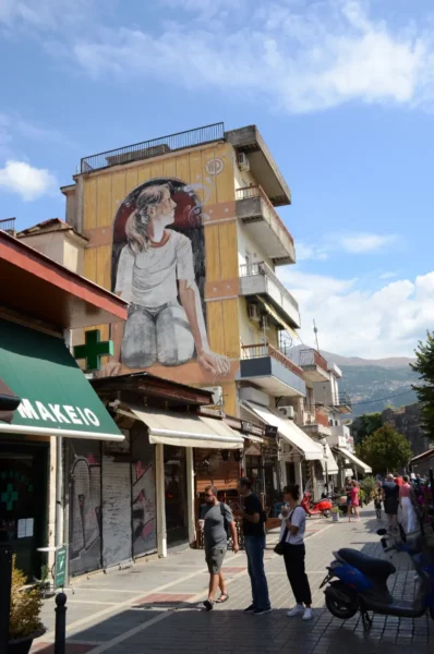 Colorful street art adorns a building in Ioannina, Greece, with pedestrians exploring local shops under a clear blue sky.