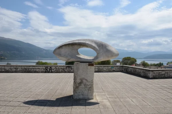 Sculptural artwork atop a stone pedestal with a lake and mountains in the background, showcasing Ioannina's serene landscape.
