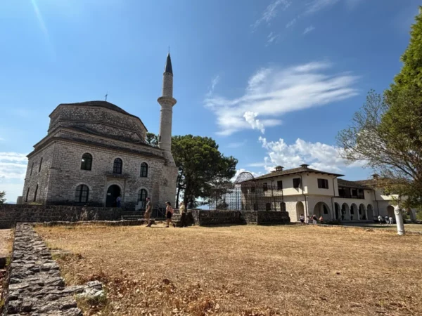Historic stone building with a minaret and surrounding greenery in Ioannina, Greece, under a clear blue sky.