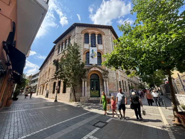 Historic stone building with vibrant greenery and pedestrians in Ioannina, Greece, under a bright blue sky.