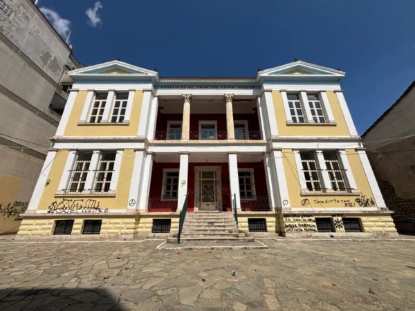 Bright yellow neoclassical building with white columns and a stone pathway, set against a clear blue sky in Ioannina, Greece.