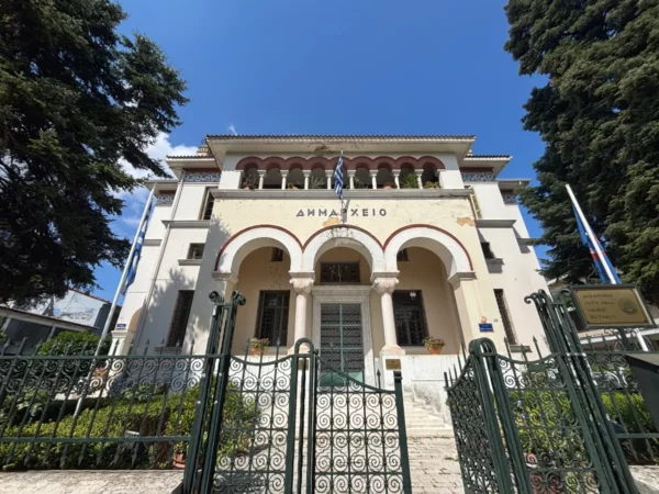 Beautifully detailed town hall with arched entrance and Greek flags, surrounded by greenery in Ioannina, Greece.