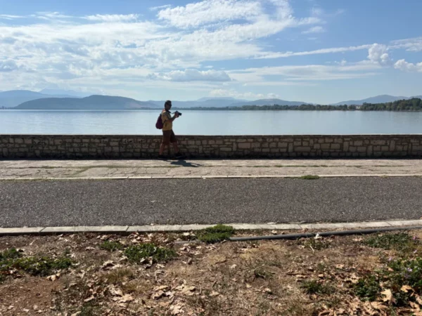 A person stands by the serene lake in Ioannina, Greece, with mountains in the background and a clear blue sky.