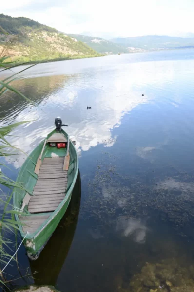 A green boat rests at the edge of the tranquil waters near Ioannina, Greece, reflecting the sky and surrounding landscape.