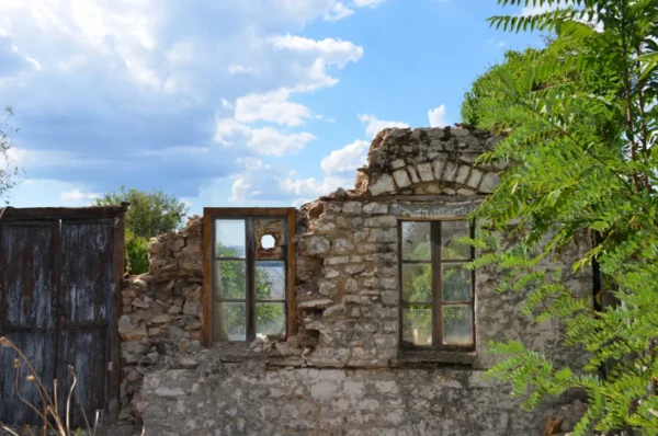 Ruined stone wall with broken windows surrounded by greenery under a bright sky on Ioannina Island, Greece.