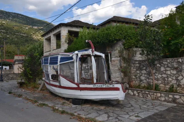 A rustic boat rests against a stone wall near traditional buildings under a cloudy sky in Ioannina, Greece.