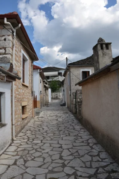Cobblestone alley lined with traditional stone houses under a blue sky dotted with clouds in Ioannina, Greece.