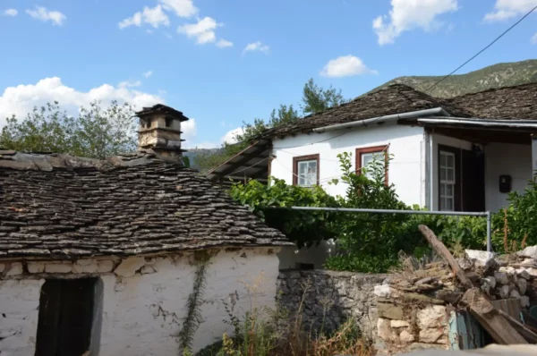 Traditional stone houses with distinctive slate roofs, surrounded by greenery in Ioannina Island, Greece. Bright blue sky adds to the scenic charm.