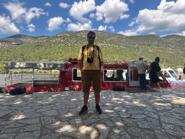 Brightly dressed traveler standing on a stone walkway near a red boat on the water in Ioannina, Greece, with mountains in the background.