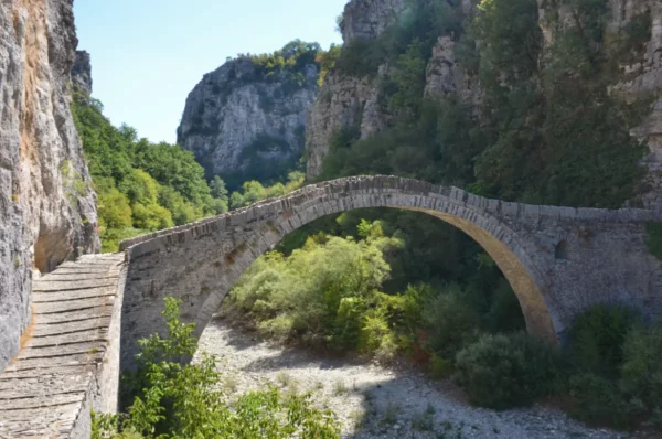 Single-arch stone Kokkoris (Noutsos) Bridge spanning the Voidomatis River in Zagorochoria, Epirus, framed by steep limestone cliffs and summer greenery.