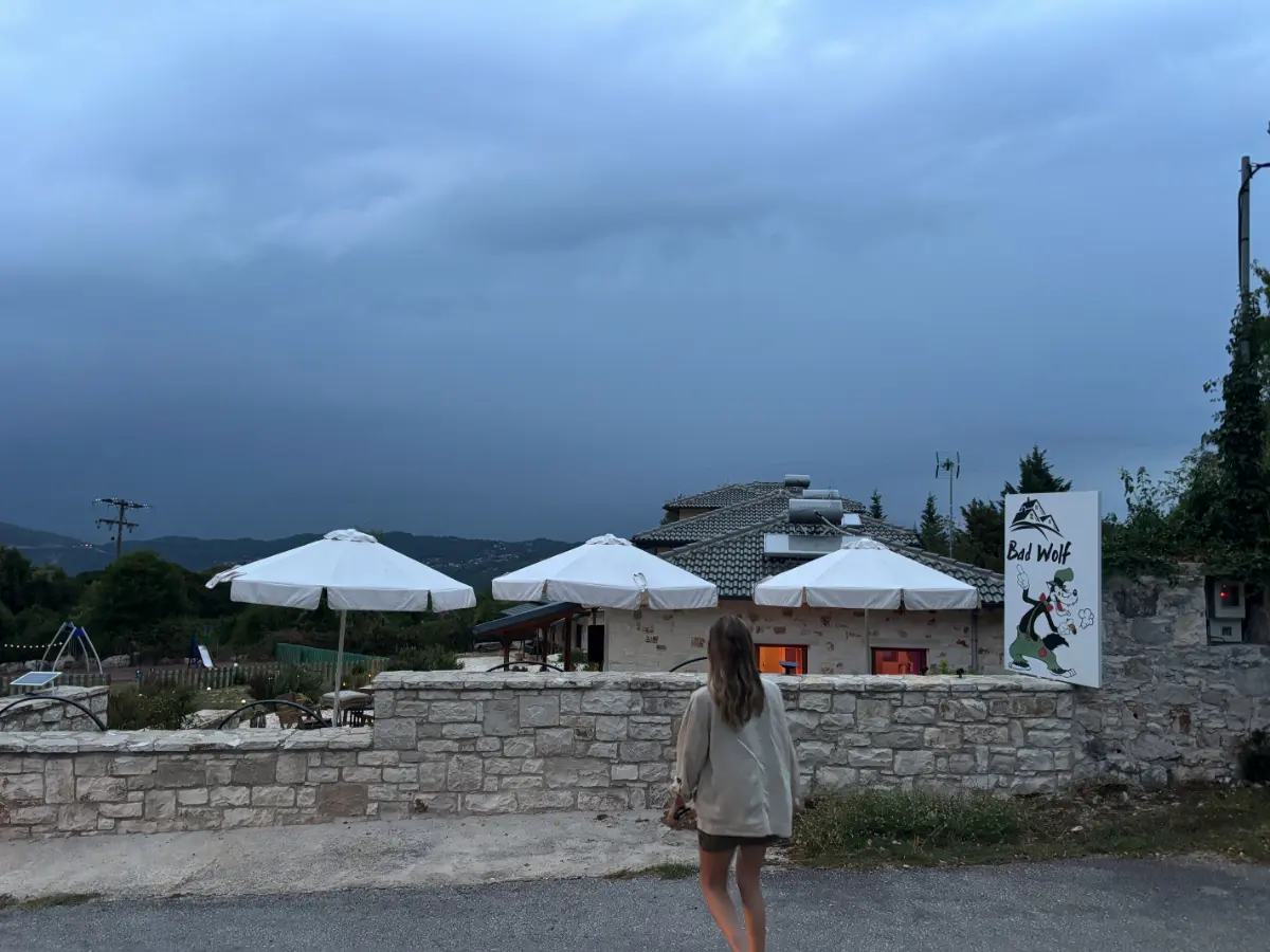 Woman walking toward Big Bad Wolf guesthouse in Kostitsi, with umbrellas and dark storm clouds above.