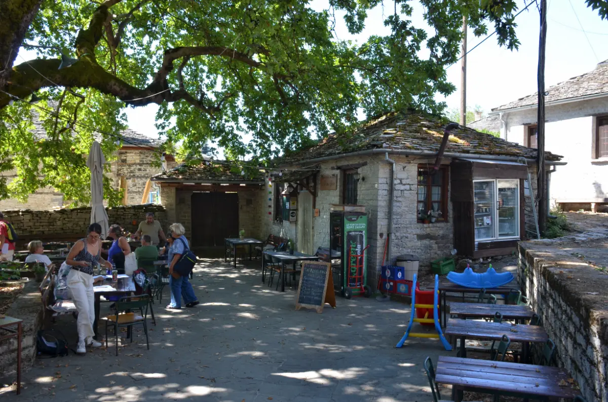 Village square in Koukouli, Zagorochoria, with a traditional stone café under a large plane tree, outdoor tables in the shade, and locals gathered on a summer afternoon.