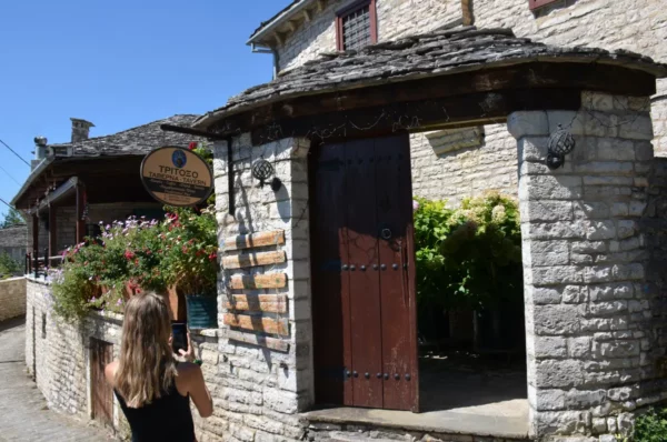 Charming stone building with a wooden entrance, colorful flowers, and a woman taking a photo in Koukouli, Zagorochoria, Greece.