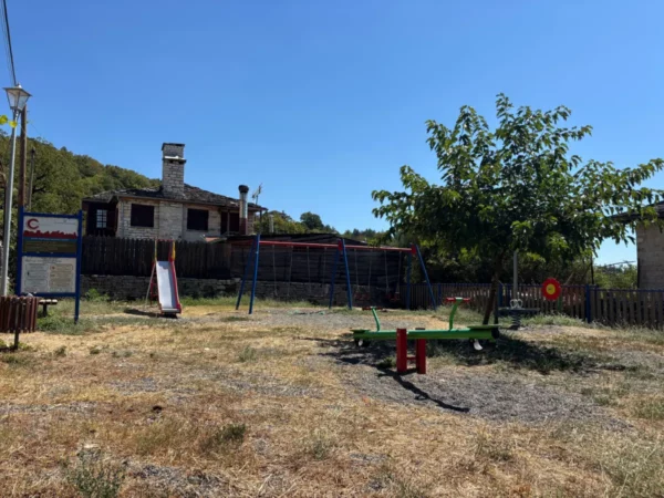 Koukouli features a playground with swings and a slide, surrounded by dry grass and traditional stone houses in Zagorochoria, Greece.
