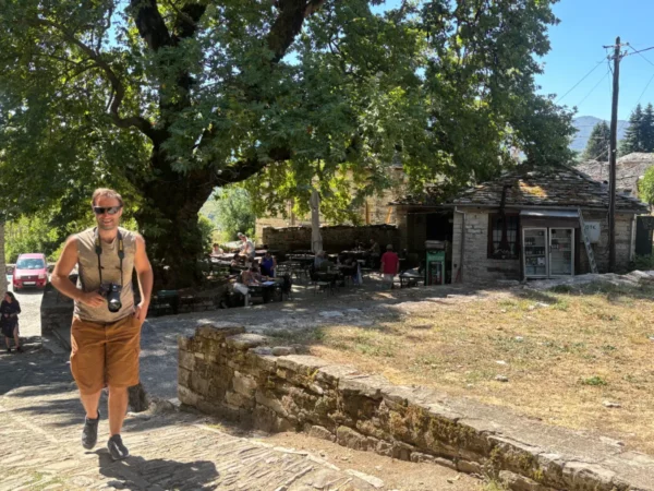 A traveler walks along a stone path in Koukouli, Zagorochoria, surrounded by greenery and traditional stone buildings.