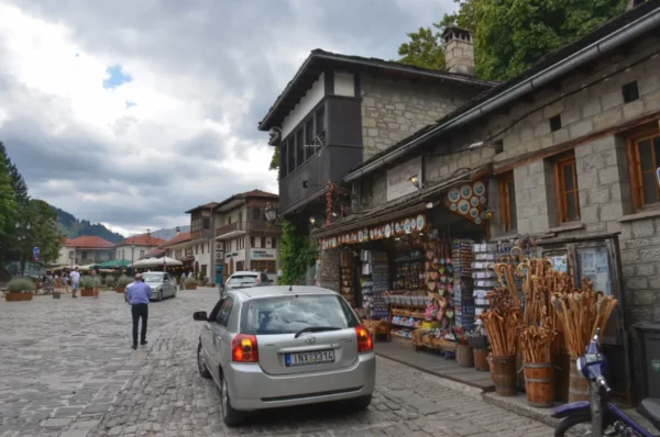 Averof Street in Metsovo, with stone-paved road, souvenir shops, cars, and mountain scenery.
