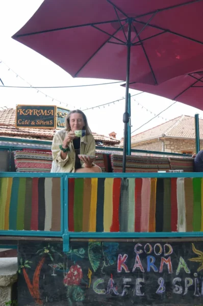 Karma Café & Spirits in Metsovo — woman sitting on a colorful balcony under umbrellas, with the café sign and rooftops behind.
