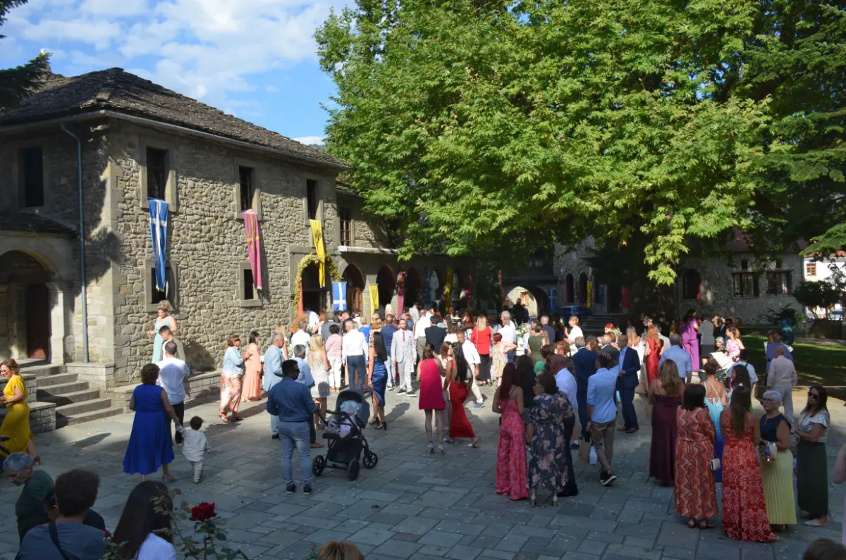 Wedding crowd outside Agia Paraskevi Church in Metsovo, gathered in the central square under tall trees in the Pindus mountains.