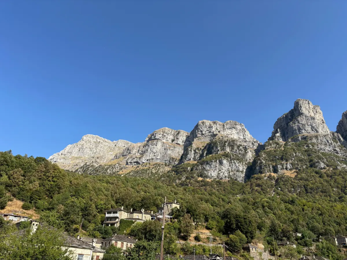 Limestone cliffs of Mount Tymfi rising above Papigo village in the Zagori region, with forested slopes and clear blue summer sky.