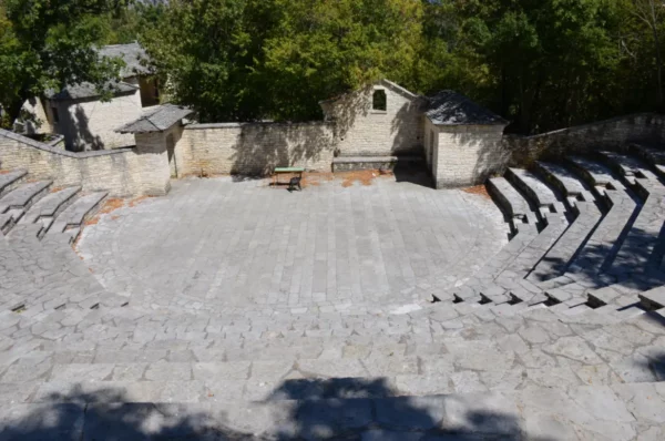 Ancient stone amphitheater with a circular stage, surrounded by lush greenery, located in Monodendri, Zagorochoria, Greece.