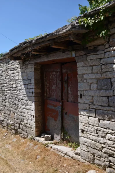 Rustic stone wall with a weathered wooden door, surrounded by natural greenery in Monodendri, Zagorochoria, Greece.