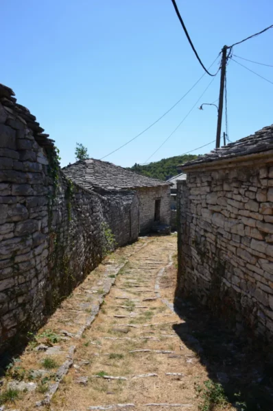 Cobblestone path lined with stone houses under a clear blue sky in Monodendri, Zagorochoria, Ioannina, Greece.